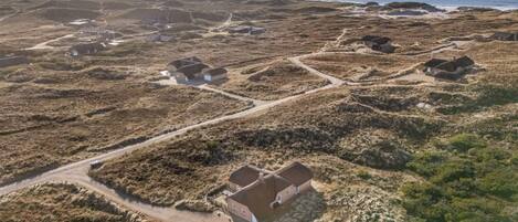 Luchtbeeld van een kustgebied met verschillende huizen, een onverharde weg en windturbines in de verte.
