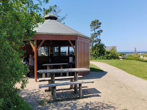 A wooden gazebo with a bench, surrounded by greenery and a clear view of the ocean.