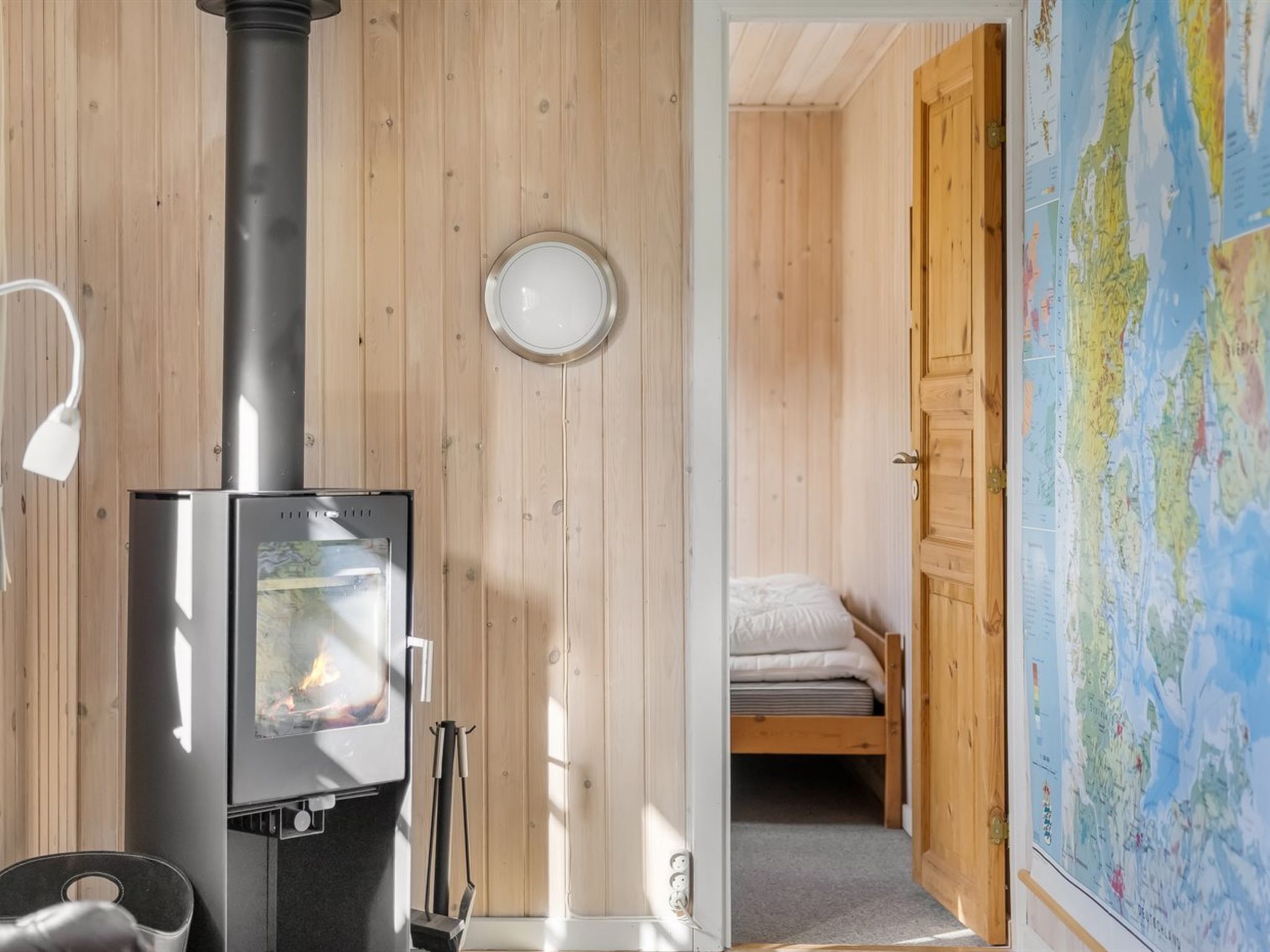 A room with a wood-burning stove, a bed with a white mattress, a map on the wall, and a wooden door.