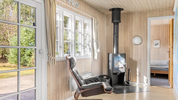 A room with a large window, a black chair, a wooden floor, and a wood-burning stove.