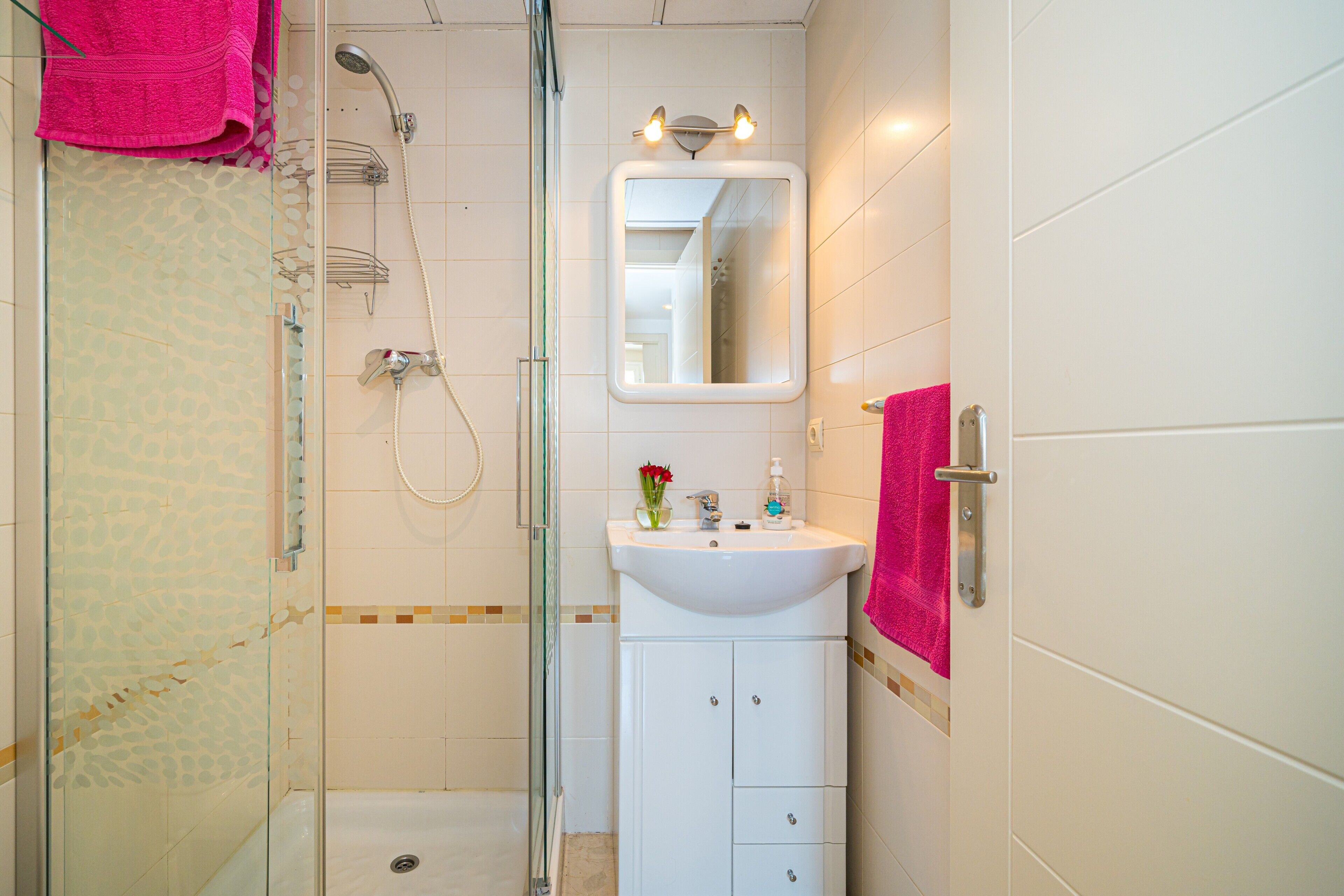 Een badkamer met een douchecabine van glas, een wit wastafelmeubel met een spiegel erboven en een roze handdoek aan de deur.