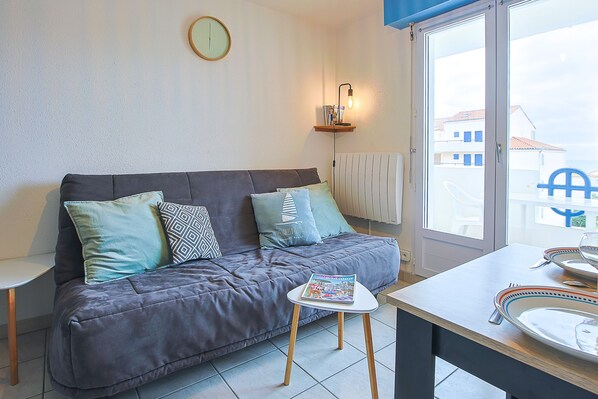 A modern living room with a grey sofa, a small white table, and a window with a view of buildings.