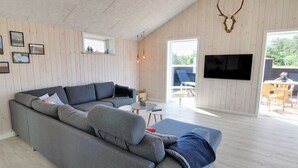 A modern living room with a grey sectional sofa, a flat-screen TV mounted on the wall, and a large window providing natural light.