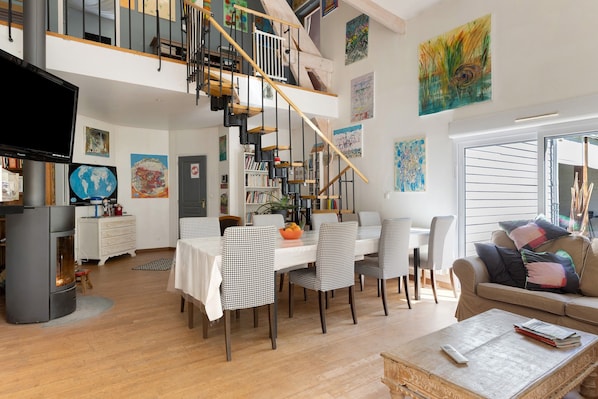 A spacious living area with a staircase, dining table, chairs, a sofa, and a television.