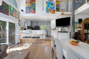 A modern kitchen with a central island, a dining area with a houndstooth patterned bench, and a living area with a flat-screen TV.