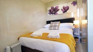 Uma cama bem arrumada com roupa de cama branca e um cobertor amarelo mostarda, uma cabeceira preta e uma obra de arte floral acima.