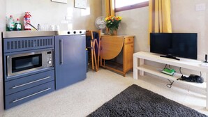 Uma cozinha compacta com micro-ondas, forno e pia. Há uma escrivaninha de madeira com um ventilador e um vaso de flores. Uma TV está sobre um suporte branco, com um controle remoto e um dispositivo verde.