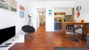 Una sala moderna con televisor de pantalla plana, una silla negra, una mesita y una zona de cocina con una isla de madera y taburetes de bar.