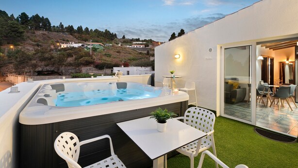 A modern outdoor hot tub area with a table and chairs, a small potted plant, and a view of a hillside. - Vacation Home "Ático Miranda" with Jacuzzi, Views of El Teide & Wi-Fi (Icod de los Vinos)