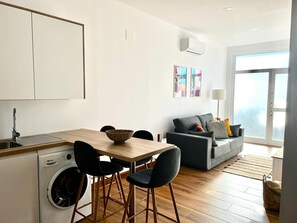 A modern kitchen with a washing machine, a dining area with a table and chairs, a gray sofa with cushions, and a large window with a view.