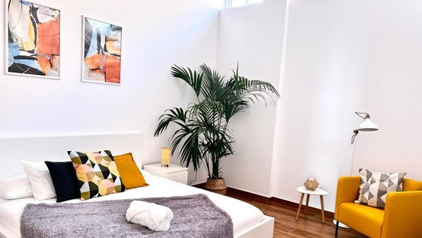 Una habitación de hotel moderna con una cama, un sillón amarillo, una planta en maceta y arte abstracto en las paredes.