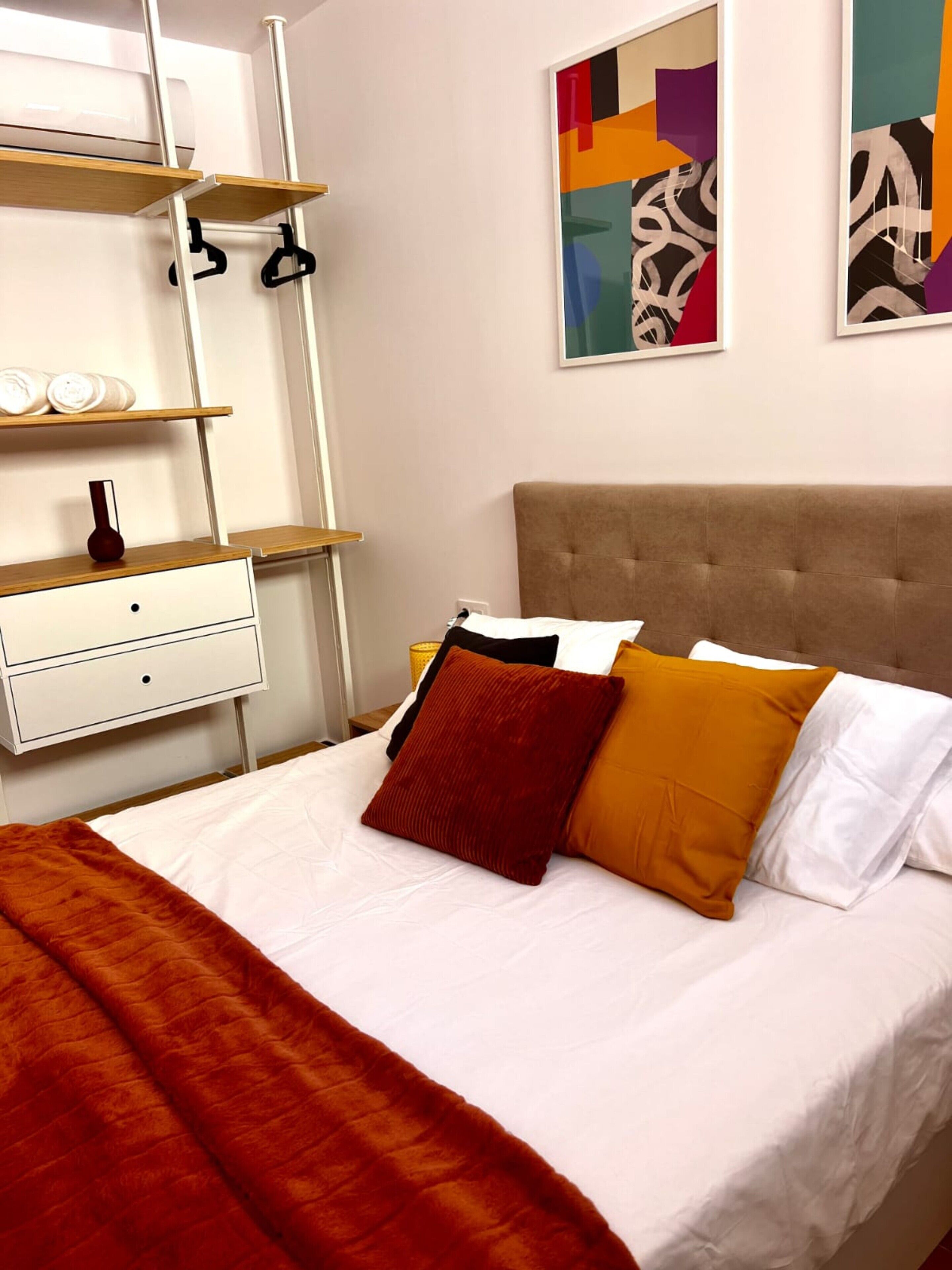 Una cama bien hecha con un cabecero marrón, una mesita de noche blanca con un florero, y arte abstracto en la pared.