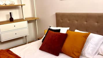 Una cama bien hecha con un cabecero marrón, una mesita de noche blanca con un florero, y arte abstracto en la pared.