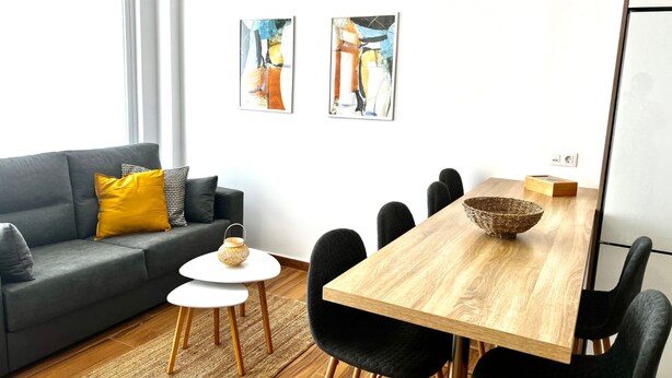 A modern living room with a grey sofa, a small round table, a wooden dining table with chairs, and two abstract paintings on the wall. - Vacation Home "Amber L5" with Sea View & Wi-Fi (Armeñime)
