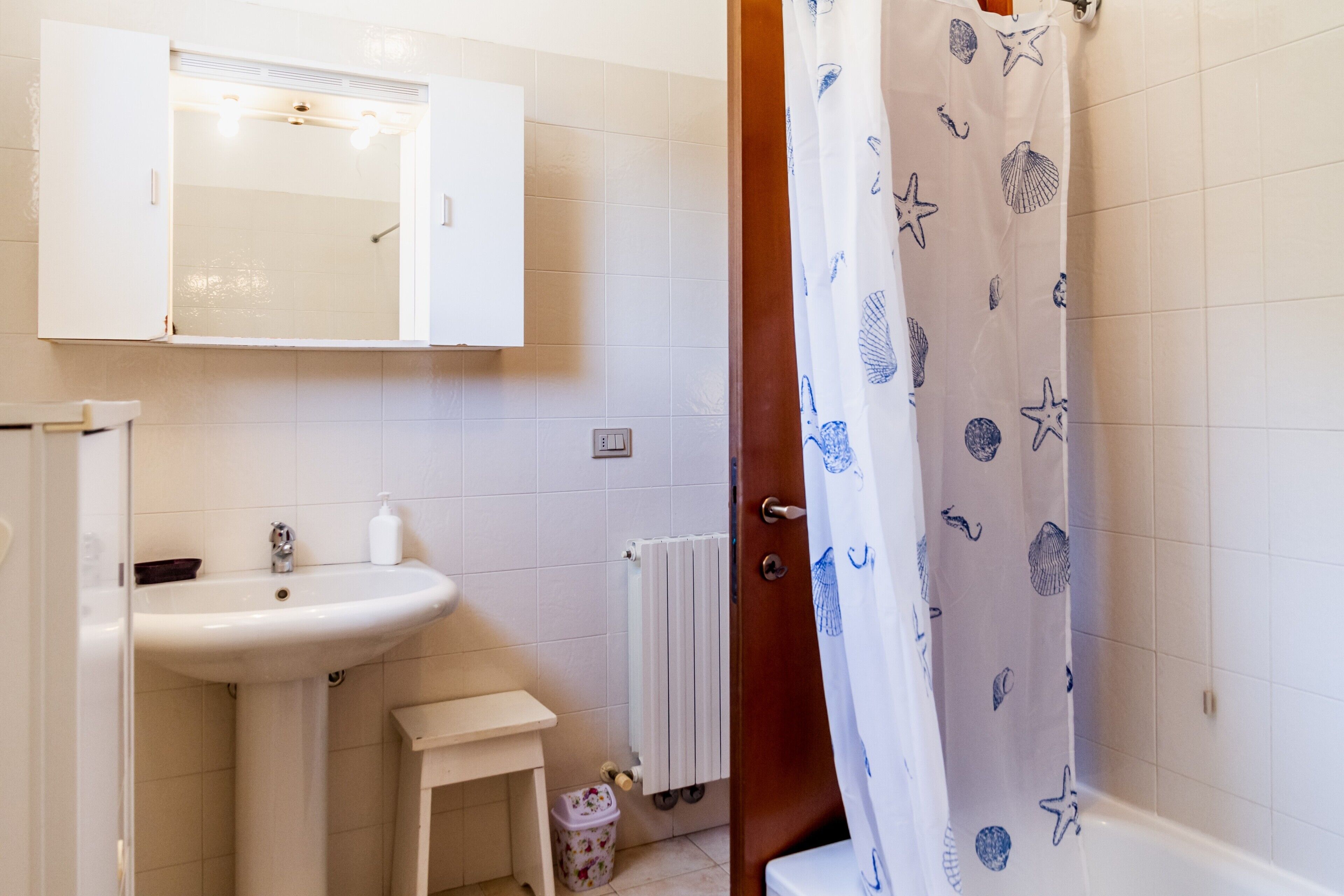 Une salle de bain avec un lavabo blanc, un miroir et un rideau de douche orné d’un motif de coquillages.