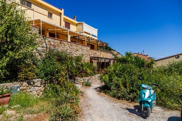 A pathway leading to a building with a stone wall and greenery, a scooter parked in front, and a clear blue sky.