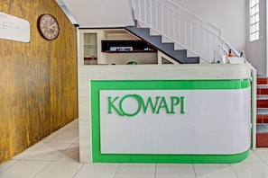 En reception med skiltet 'KOWAPI' i grøn skrift, en trævæg, et ur og en trappe i baggrunden.