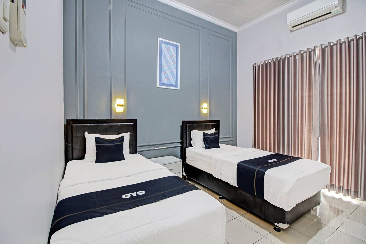 Habitación de hotel con dos camas, un ventanal con cortinas, un equipo de aire acondicionado y un cuadro en la pared.