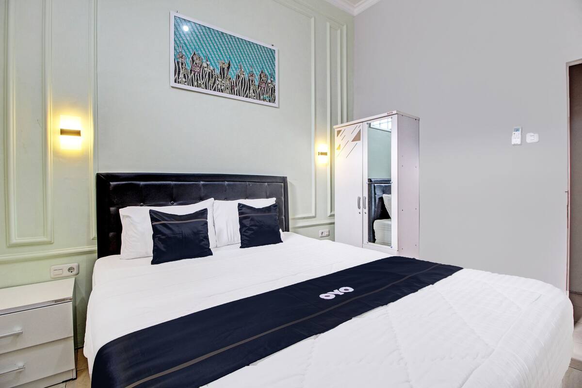 Una cama bien hecha con colcha blanco y negro y un logo, una mesita de noche blanca, una lámpara de pared y una puerta con espejo.