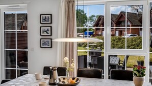 Un comedor con una mesa dispuesta para dos, un jarrón con flores y vistas a casas a través de amplias ventanas.