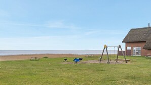 Un área con césped, un columpio, una construcción con techo de paja y vistas al mar.
