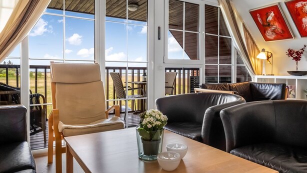 Una habitación amplia con un gran ventanal corredera que da a un balcón, una mesa de madera con un florero y un sofá de cuero negro.