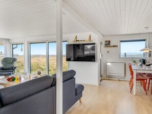 A spacious living area with a sofa, a dining table with chairs, a kitchen area with a TV, and large windows offering a view of the landscape.