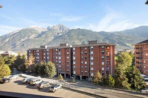 Un edificio de varios pisos con un estacionamiento al frente, rodeado de árboles y montañas al fondo.