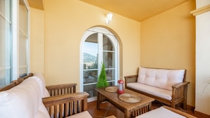 A sunlit patio with a wooden table, two cushioned chairs, a small potted plant, and a view through a large arched window. - Apartment 'Señorio De Aloha In Puerto Banus' with Mountain View, Wi-Fi and Air Conditioning (Nueva Andalucia Marbella)