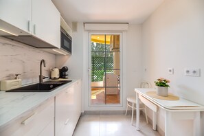 Een kleine keuken met een spoelbak, fornuis en magnetron. Er is een eethoek met een tafel en stoelen, en een schuifpui naar een terras.