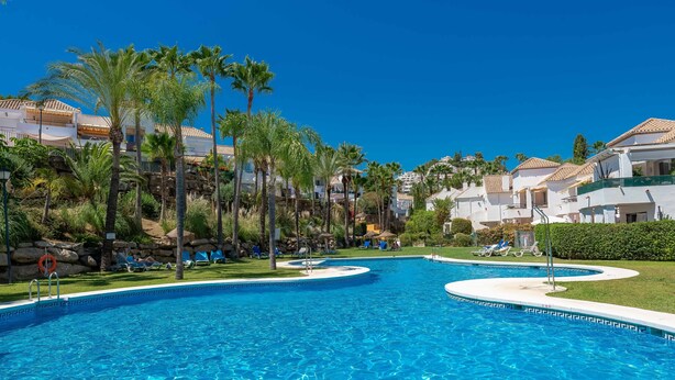 A swimming pool with a clear blue water, surrounded by palm trees and lounge chairs, with buildings in the background. - Apartment 'Señorio De Gonzaga In Marbella' with Shared Pool, Wi-Fi and Air Conditioning (Marbella)