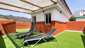 A building with a white and orange facade, a covered patio with artificial grass, and two lounge chairs. - Vacation Home "Las Monarcas" with Mountain View, Private Terrace & Wi-Fi (Teror)