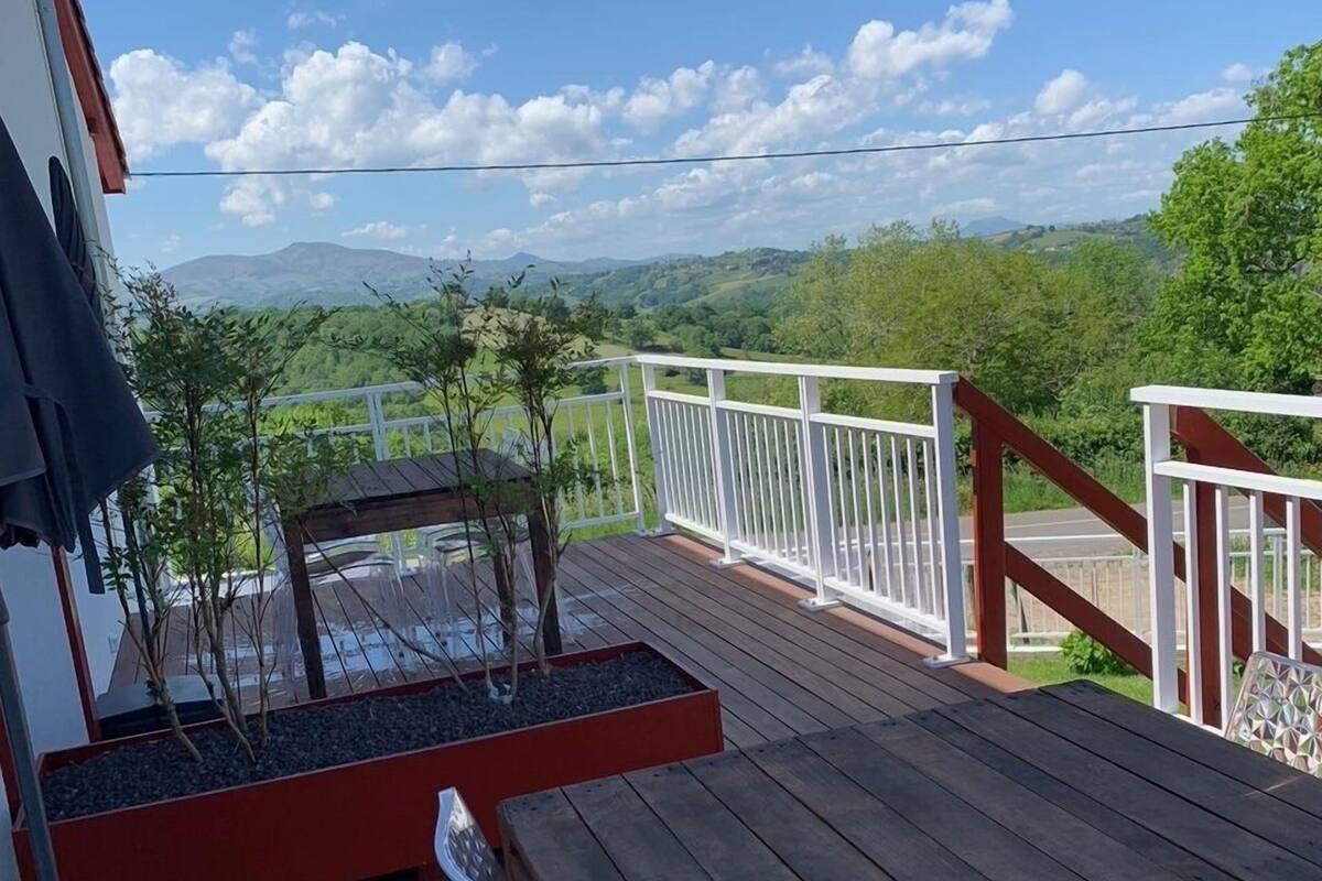Une terrasse en bois avec une balustrade blanche, un petit jardinier avec des plantes, et une vue sur des collines verdoyantes et des montagnes en arrière-plan.