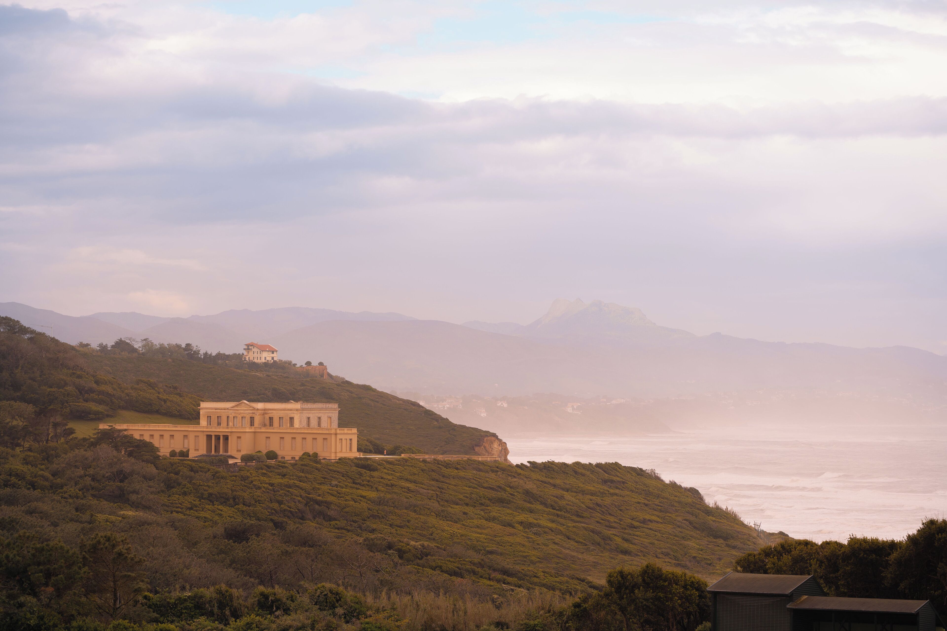 A large, historic building with a symmetrical facade, situated on a hillside overlooking a coastal area.