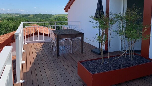 Eine Holzterrasse mit Tisch und Stühlen, ein kleiner Pflanzkasten mit Bäumen und Blick auf die Landschaft.