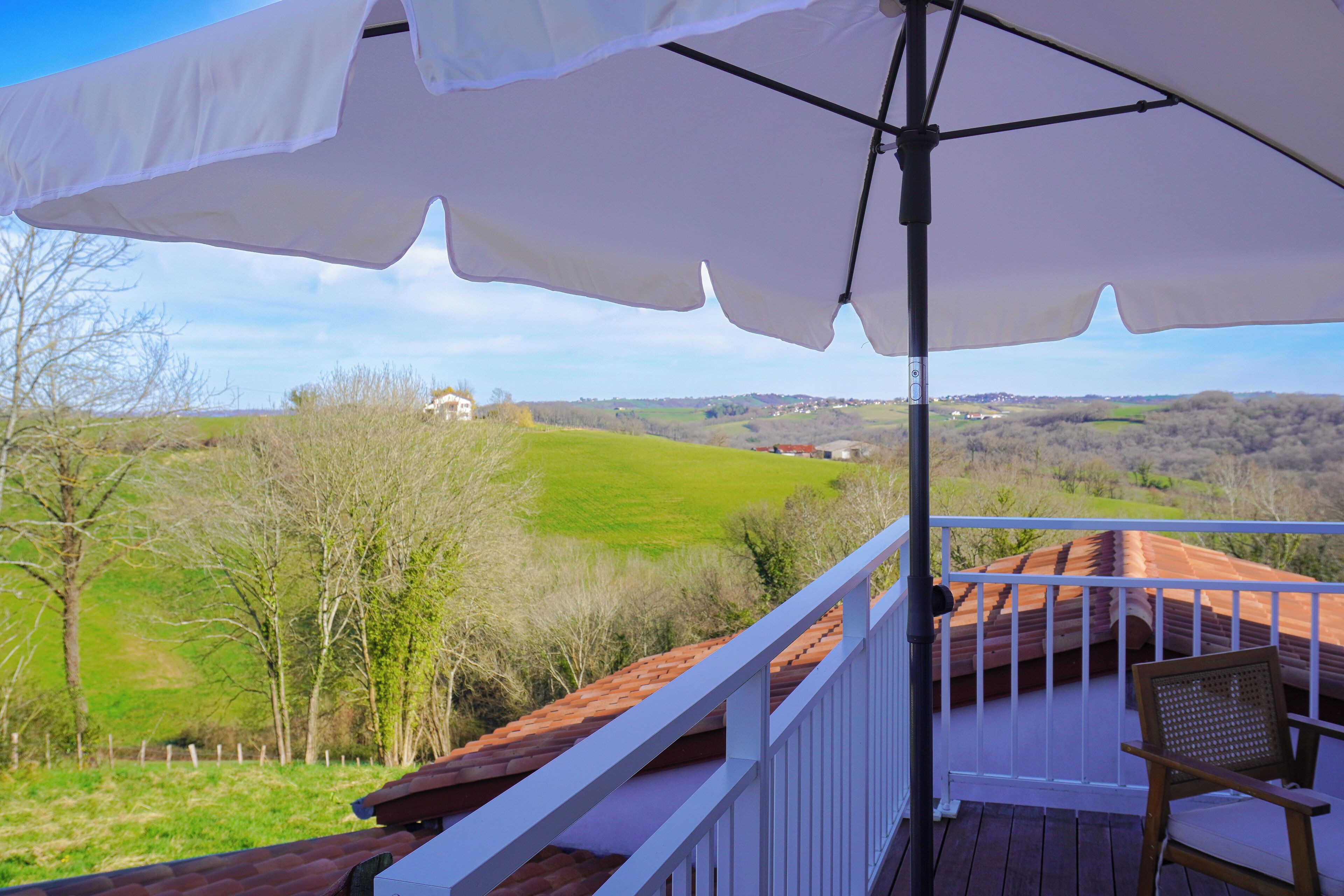 Ein Balkon mit weißem Sonnenschirm, einem Holzstuhl und Blick auf grüne Felder sowie ein weiter abgelegenes Gebäude.
