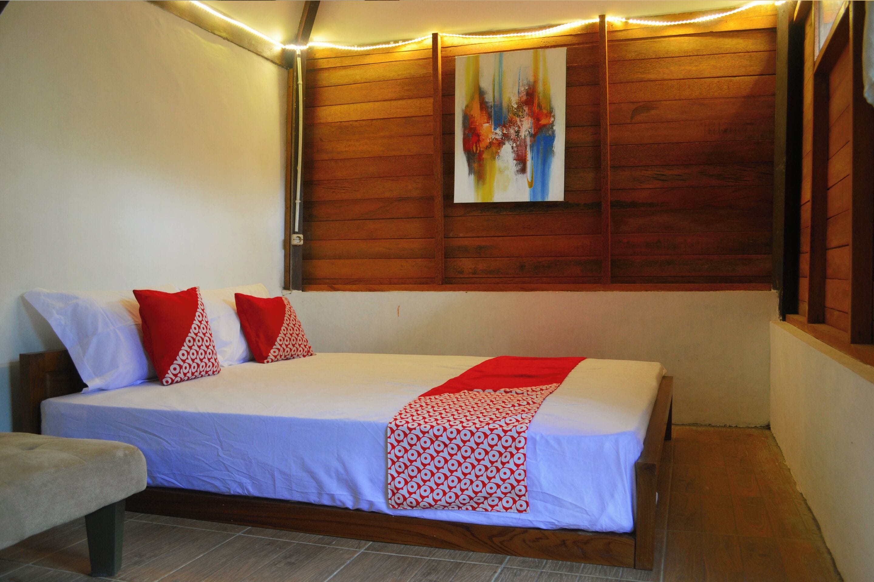 Un letto rifatto con cura, coperta bianca e rossa a motivi decorativi, una testiera in legno e un quadro astratto appeso al muro.