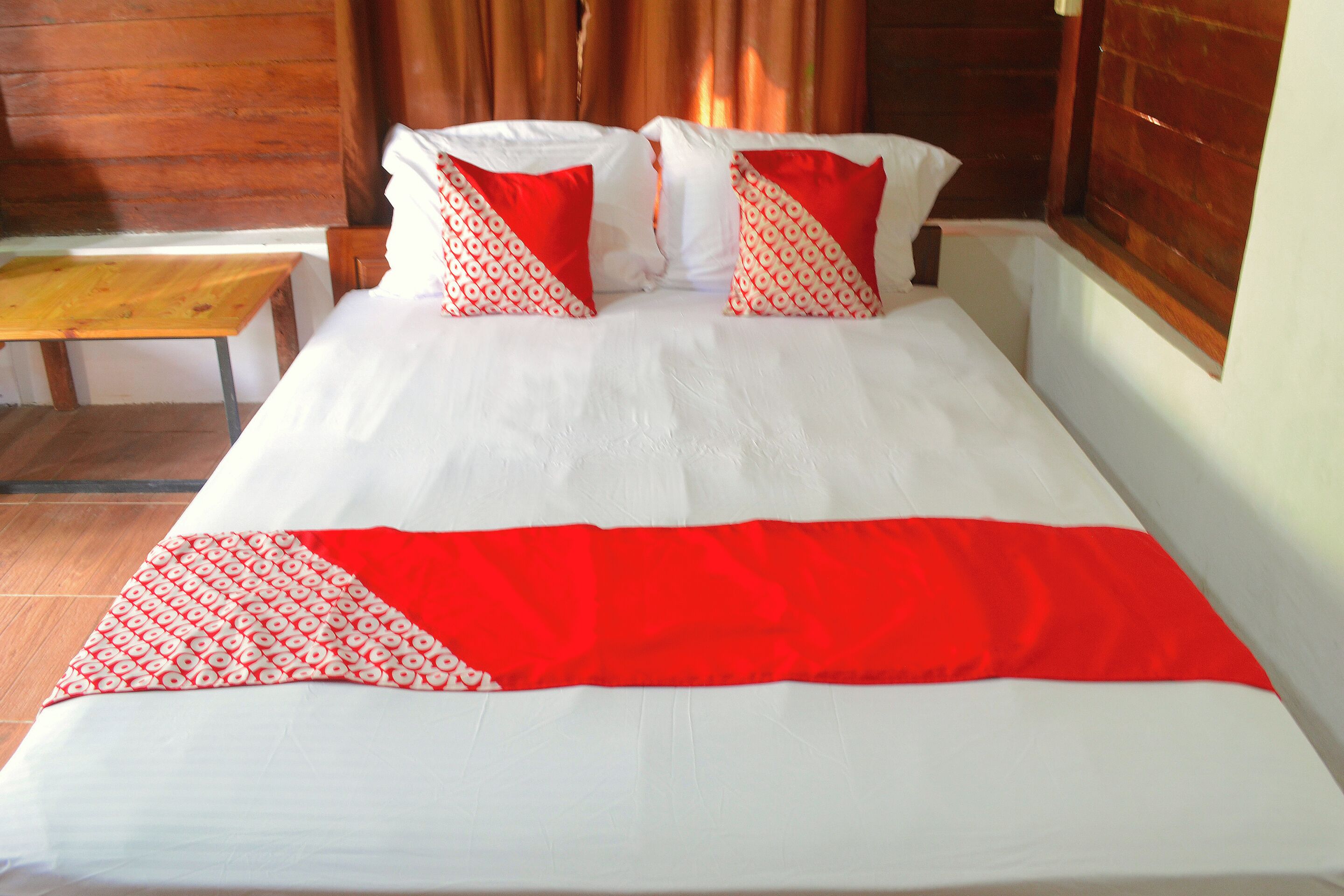 A bed with white bedding and red and white patterned pillows, a wooden headboard, and a wooden bench beside the bed.