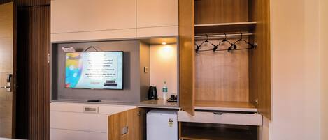 A compact hotel room with a bed, a small kitchenette, a TV, and a wooden wardrobe with hanging space.