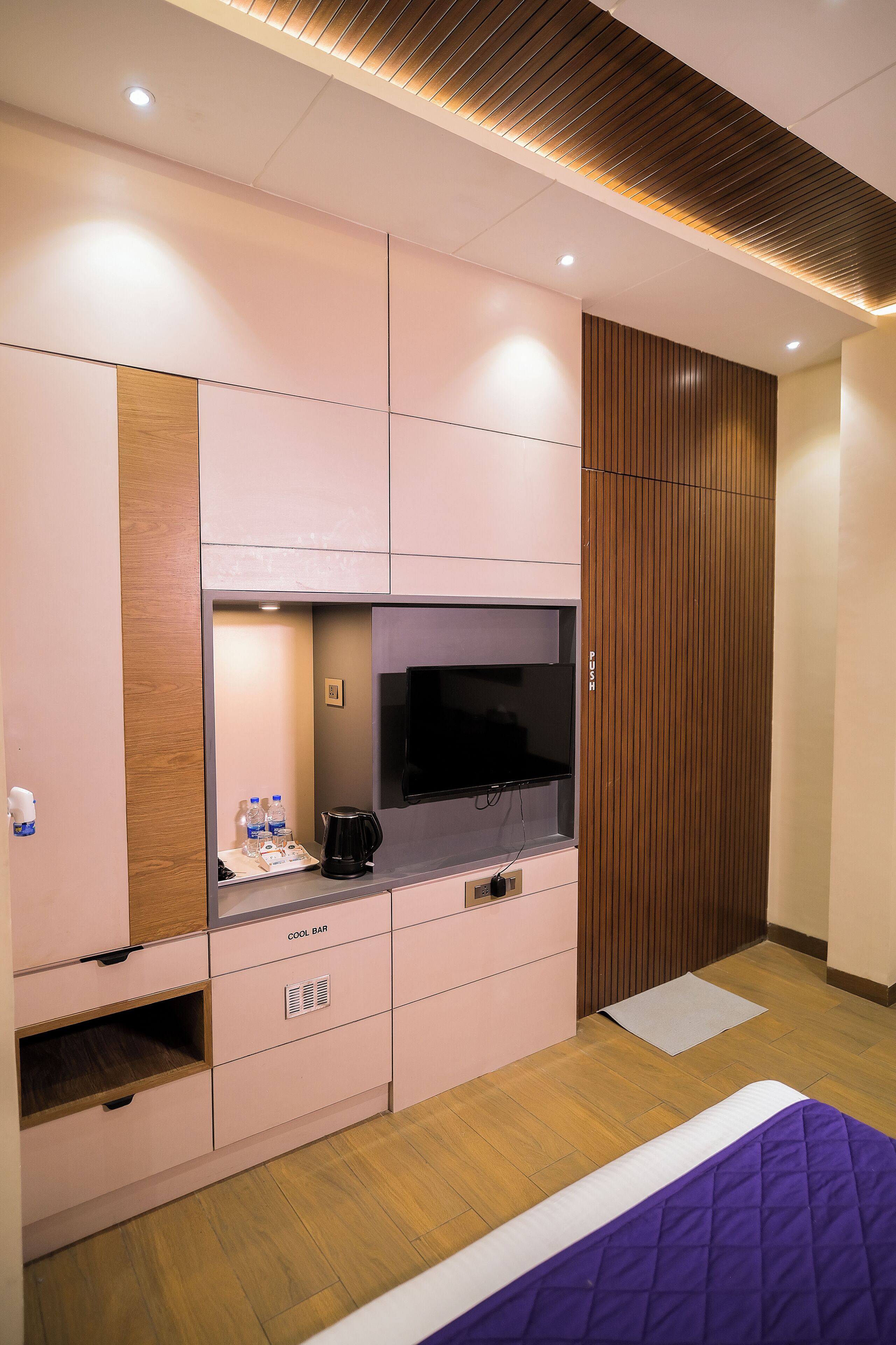 A bedroom with a bed, a TV, a coffee maker, and a shelf with water bottles.