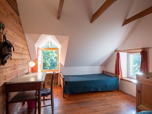 A cozy attic bedroom with a wooden floor, a desk with a lamp, a bed with a blue bedspread, and a window with curtains. - Le Petit Poirier by Interhome (Fouesnant)