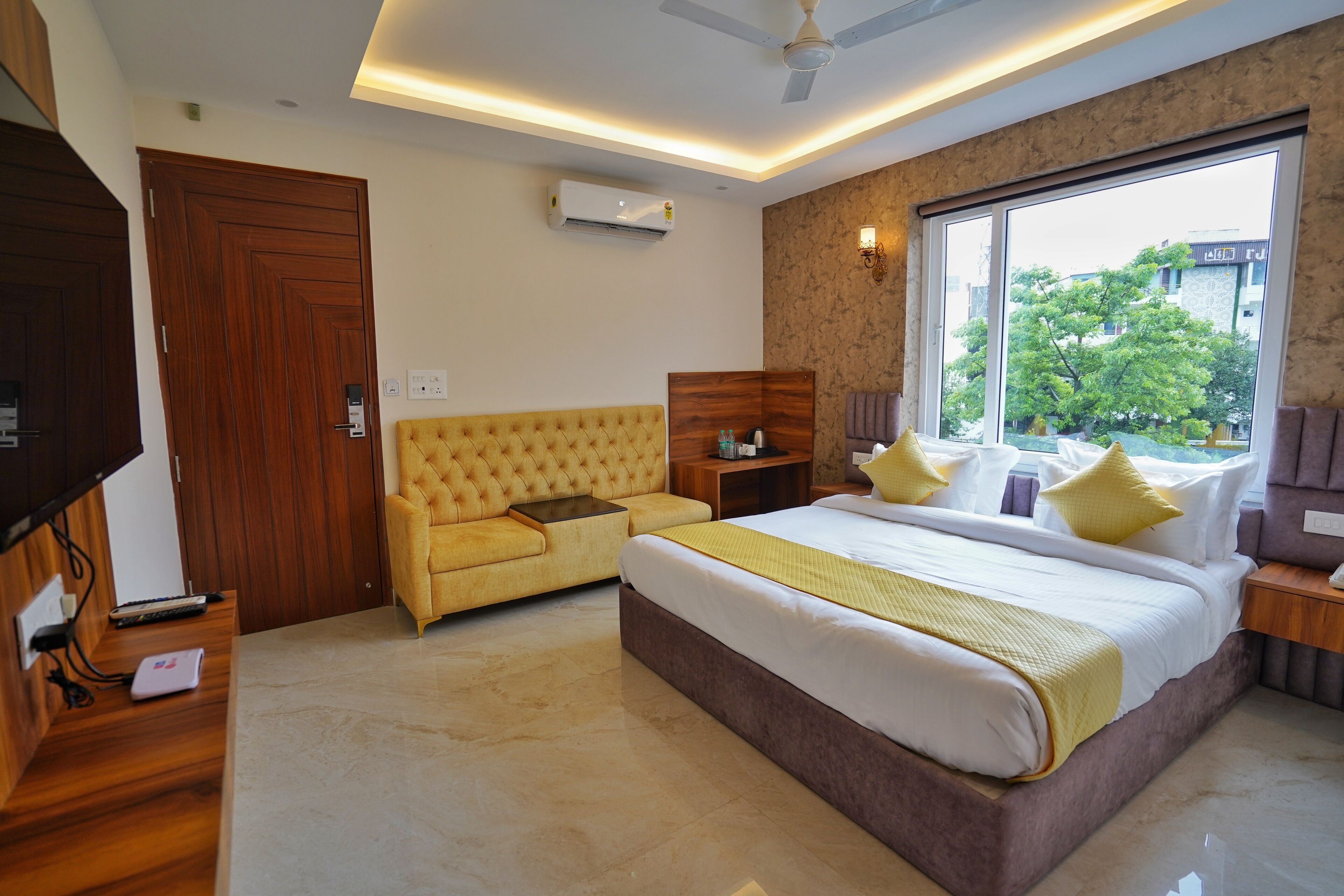 A bedroom with a large bed, a sofa, a TV, and a window with a view of trees.