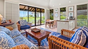 A spacious living area with a large sofa, a coffee table, and wicker furniture. There is a ceiling fan, a dining table with chairs, and a kitchen area with a bar. - Ground-Floor Retreat W/Ocean Views-Kiahuna 2166 (Koloa)