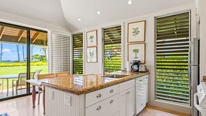 A modern kitchen with a large island, white cabinetry, and a view of a lush garden through the sliding glass doors. - Ground-Floor Retreat W/Ocean Views-Kiahuna 2166 (Koloa)