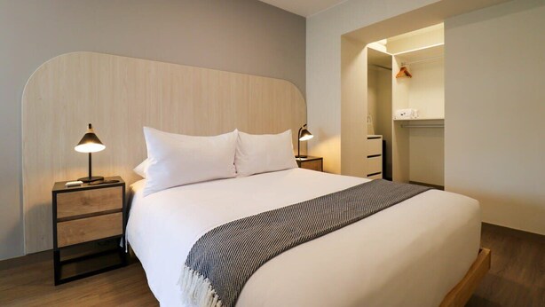 Une chambre d’hôtel moderne dotée d’un grand lit, de deux tables de chevet avec lampes, d’une tête de lit en bois et d’un dressing.