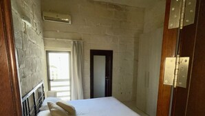 A small, single-bed room with a wooden headboard, a window with curtains, and a door with a mirror.