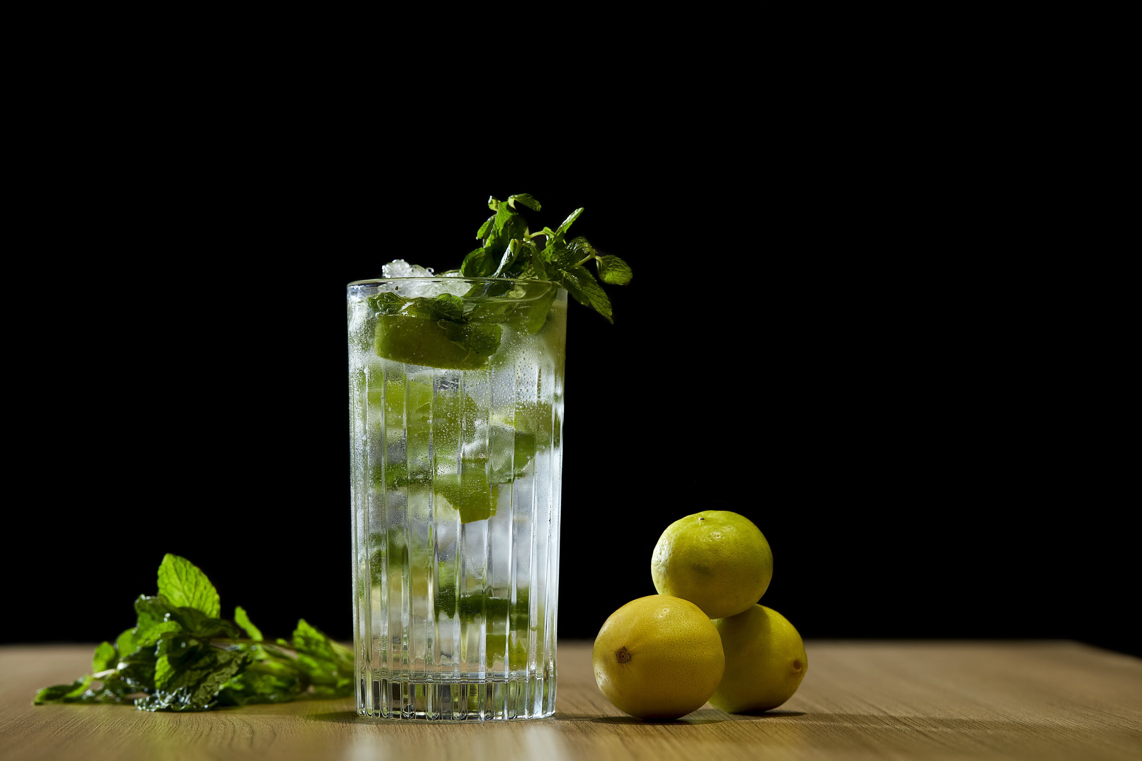 A tall glass filled with a clear liquid, ice cubes, and mint leaves, accompanied by two lemons on a wooden surface against a dark background.