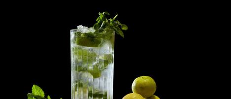 Un vaso alto lleno de un líquido transparente, cubitos de hielo y hojas de menta, acompañado de dos limones sobre una superficie de madera contra un fondo oscuro.