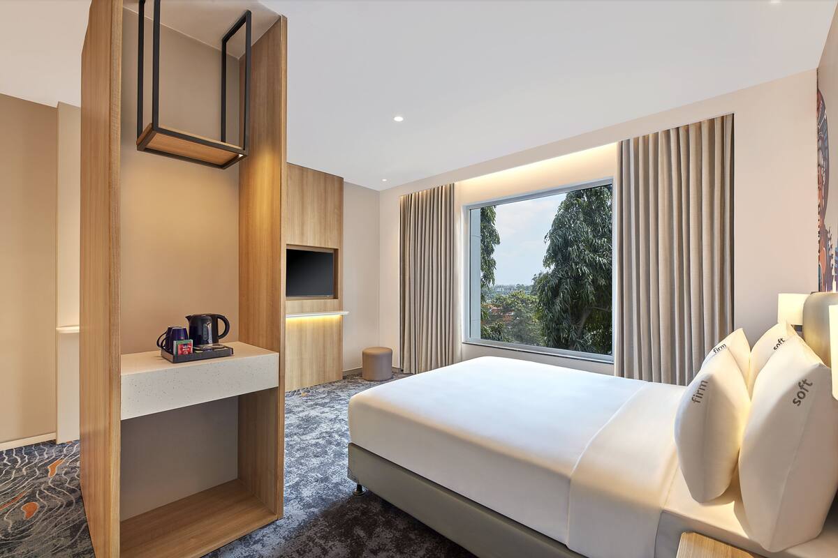 A modern hotel room with a large bed, a TV, a coffee maker, and a view of trees through the window.