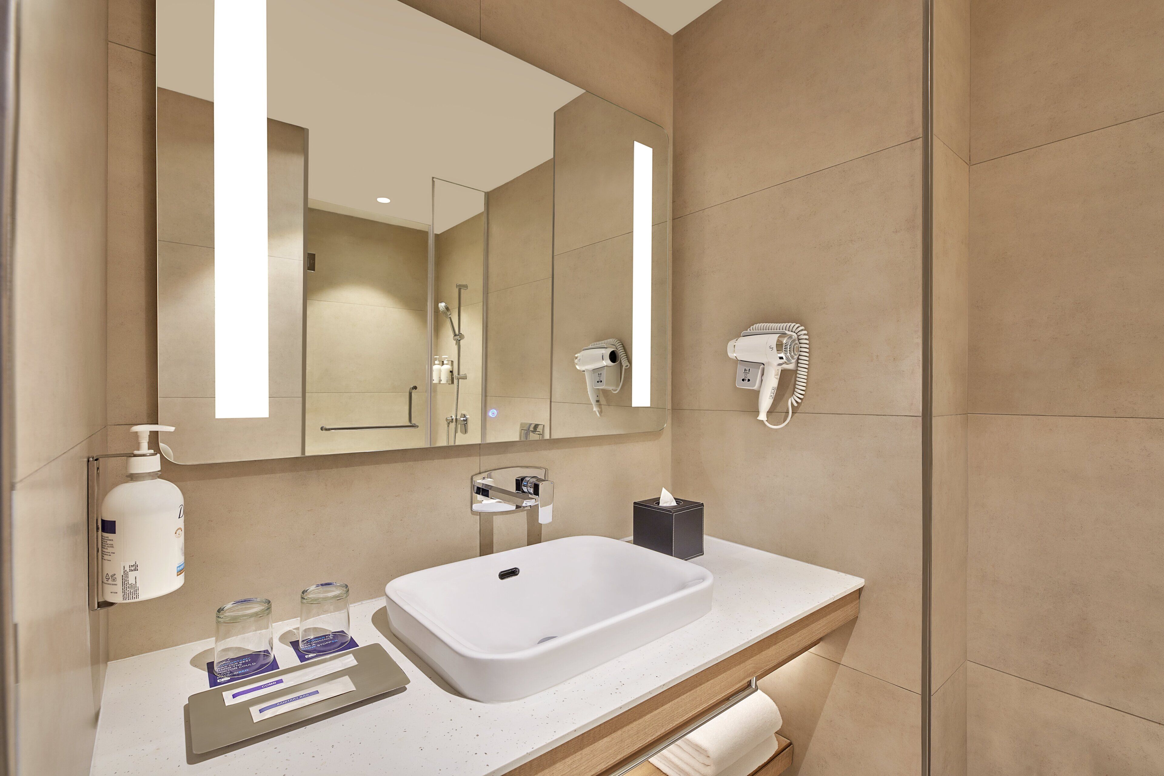 A modern bathroom with a large mirror, a white sink, a hairdryer, and a bottle of hand soap.
